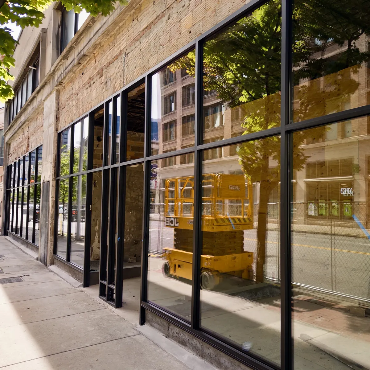Ground-floor commercial space under construction, with new black-framed storefront glass installed along the sidewalk and a yellow scissor lift reflected in the panes.